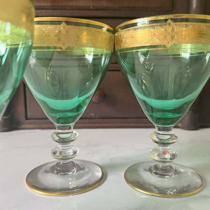 Two green glass goblets with gold trim and etched detailing on the rims, displayed on a white surface with a dark wooden background.