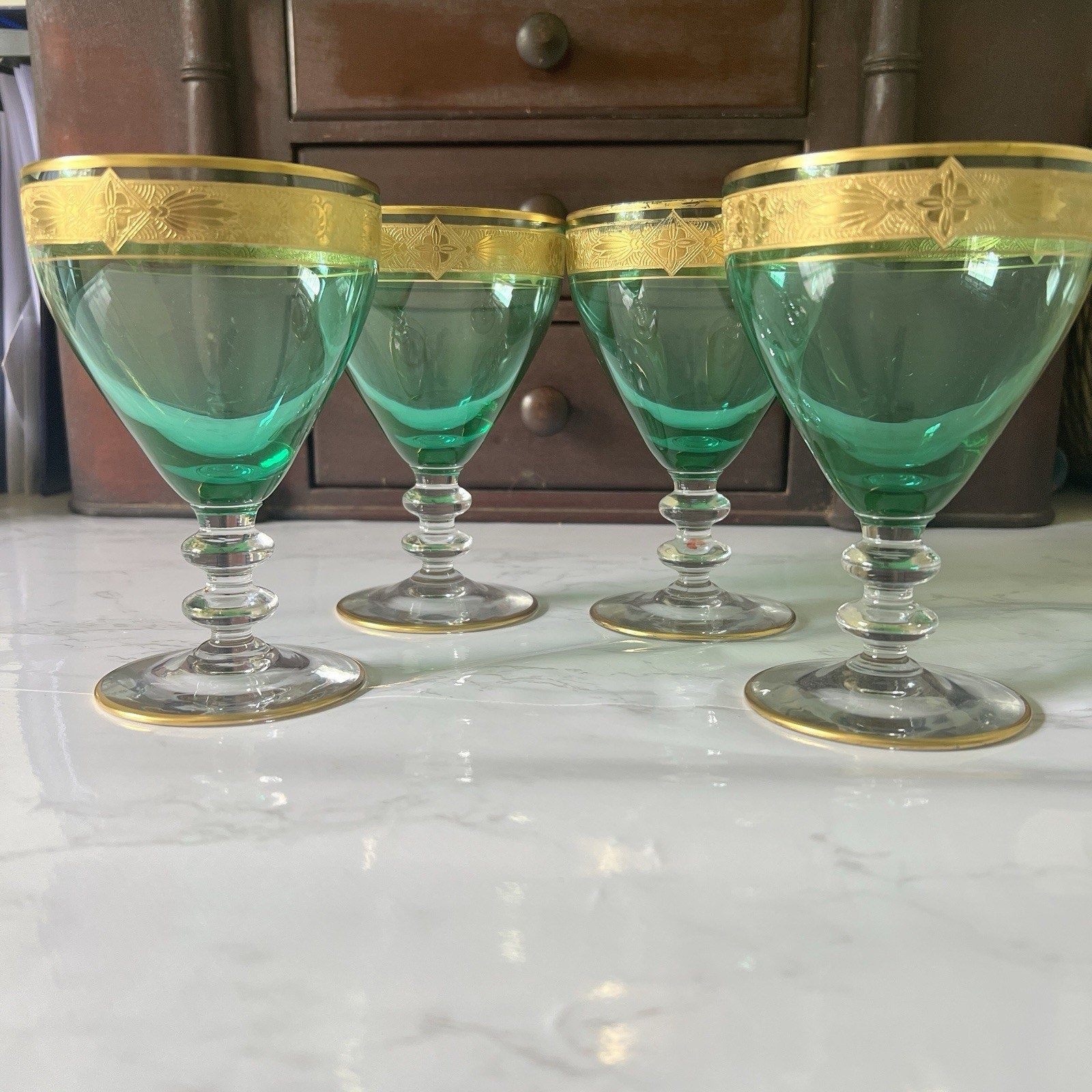 Four green glass goblets with gold decorative rims, arranged on a white marble surface in front of a dark wooden cabinet.