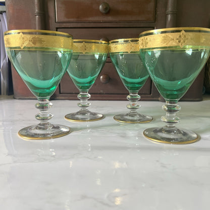 Four green glass goblets with gold decorative rims, arranged on a white marble surface in front of a dark wooden cabinet.