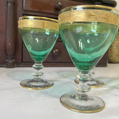 Three vintage green glass goblets with gold bands around the rims, displayed on a light marble surface with a dark wooden cabinet in the background.
