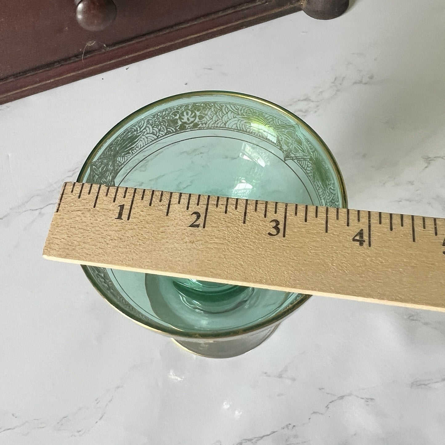 A green glass with a gold rim and floral pattern, shown with a wooden ruler to indicate its size on a white marble surface.