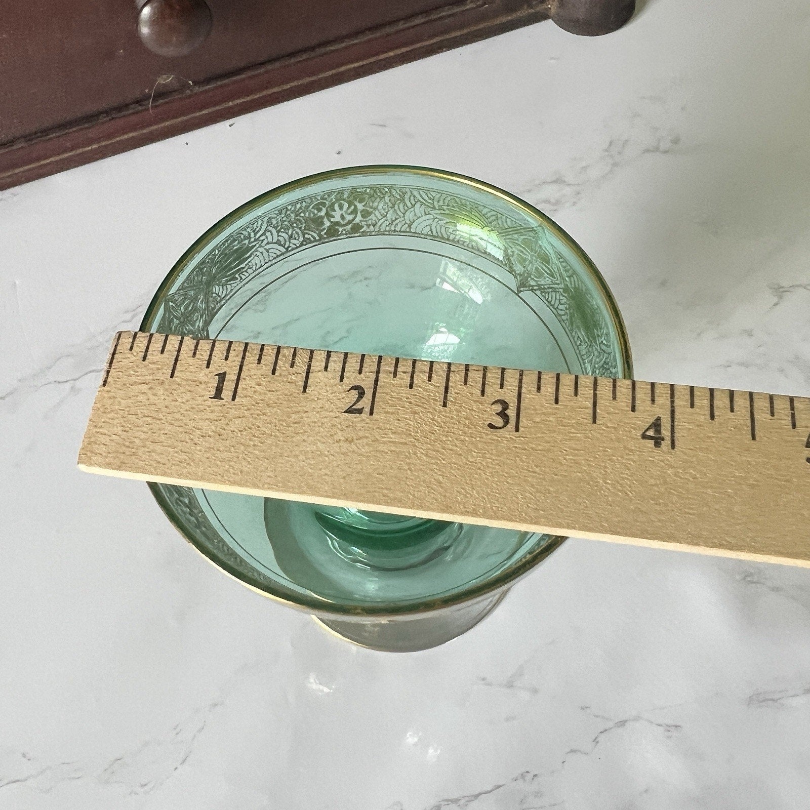 A green glass with a gold rim and floral pattern, shown with a wooden ruler to indicate its size on a white marble surface.