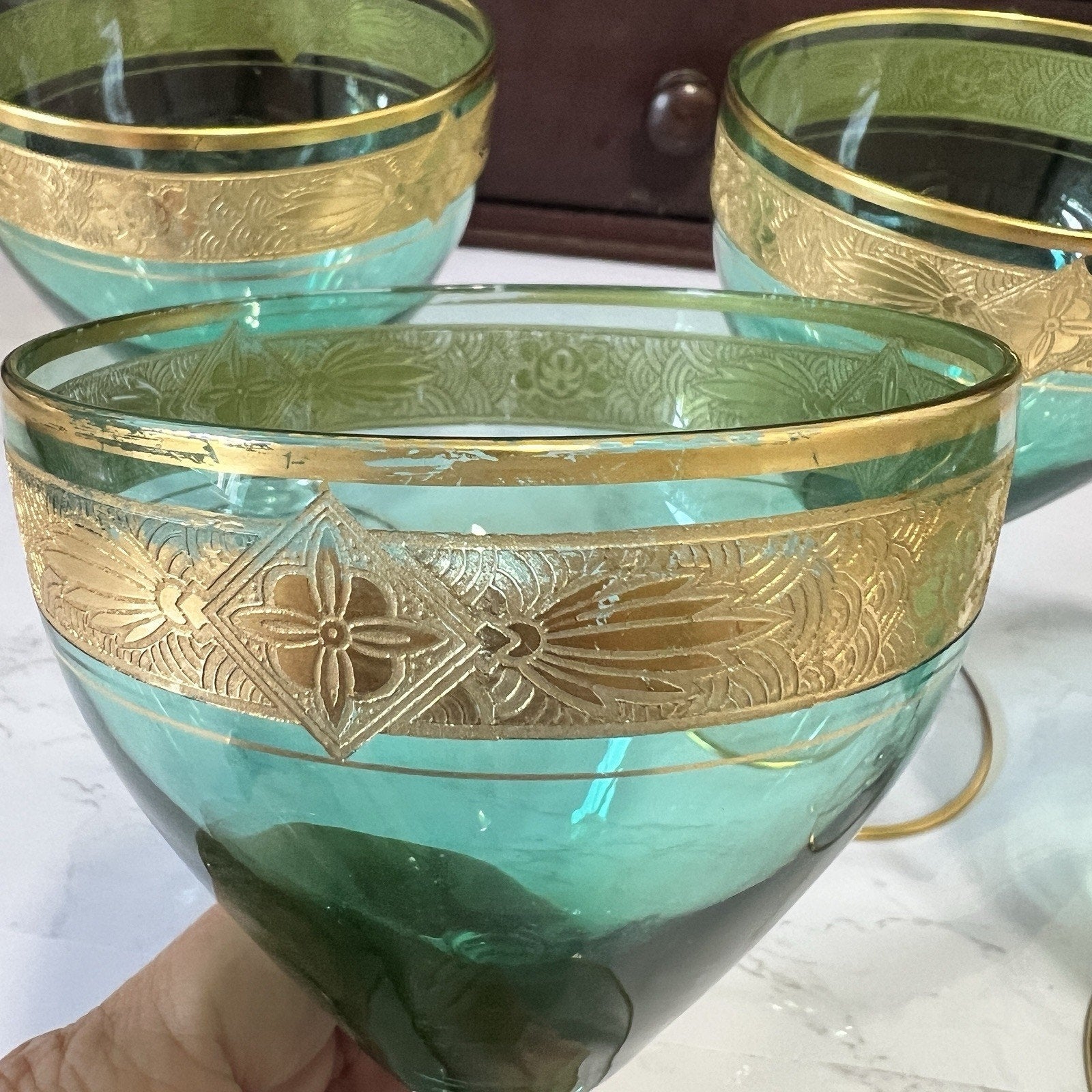 A vintage-style green glass bowl with a wide gold band featuring embossed floral and geometric patterns, held by a hand against a marble surface with other similar bowls in the background.