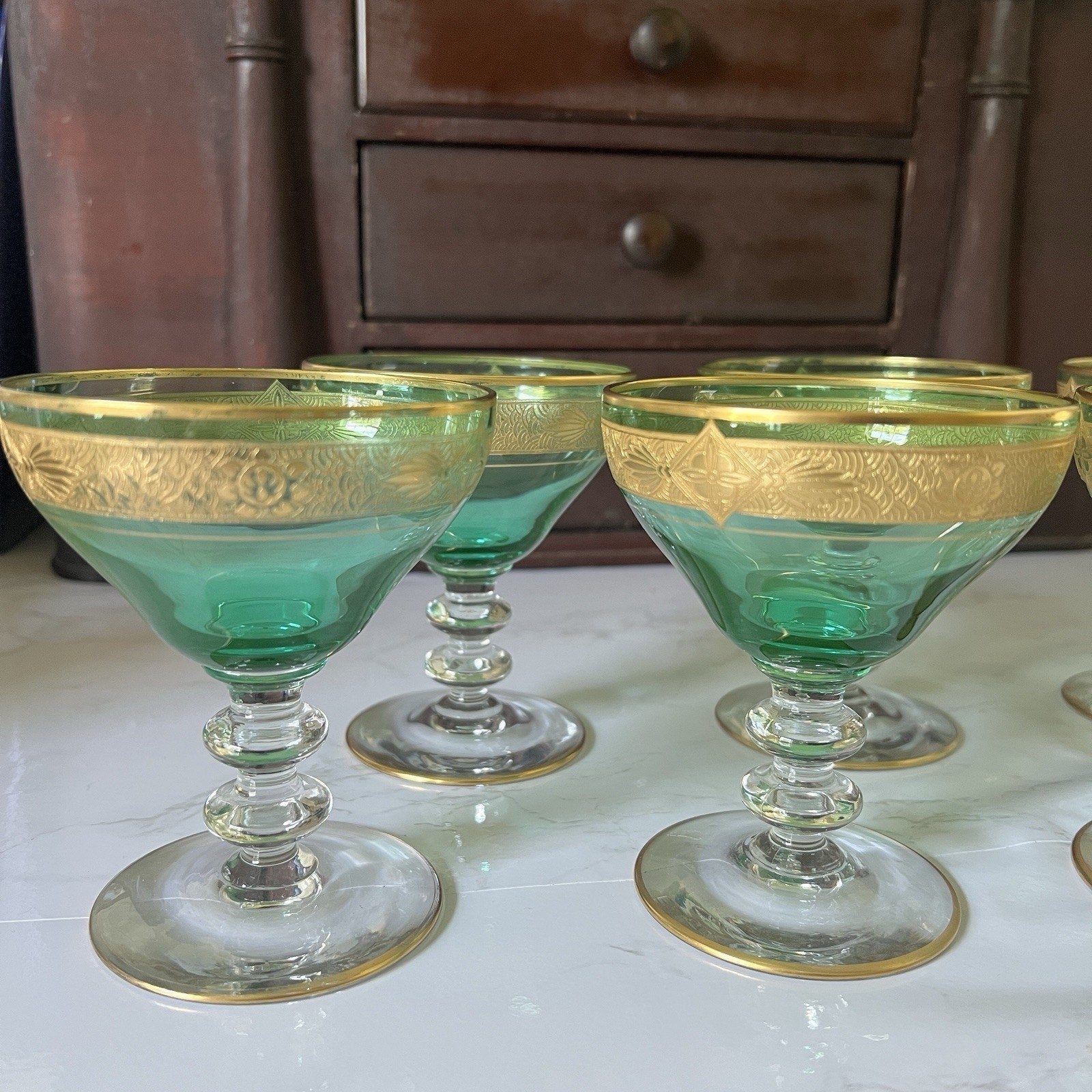 A set of vintage green and gold coupe glasses arranged on a white surface with a dark wooden cabinet in the background.