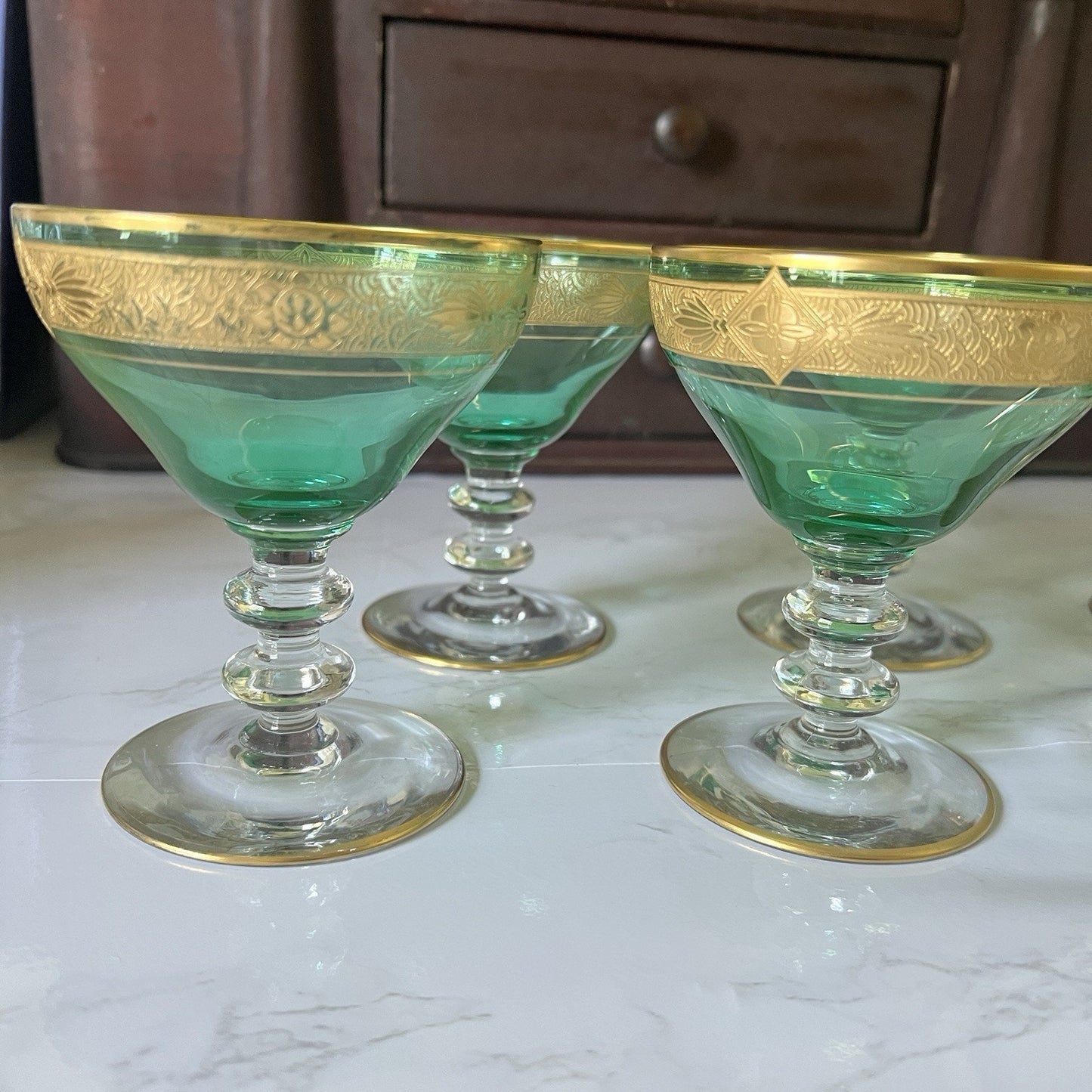 Three green glass coupe cups with gold trim and etched floral detailing near the rim, arranged on a light marble surface with a dark wooden cabinet in the background.