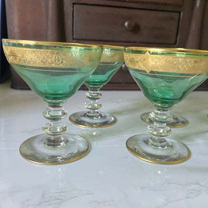 Three green glass coupe cups with gold trim and etched floral detailing near the rim, arranged on a light marble surface with a dark wooden cabinet in the background.