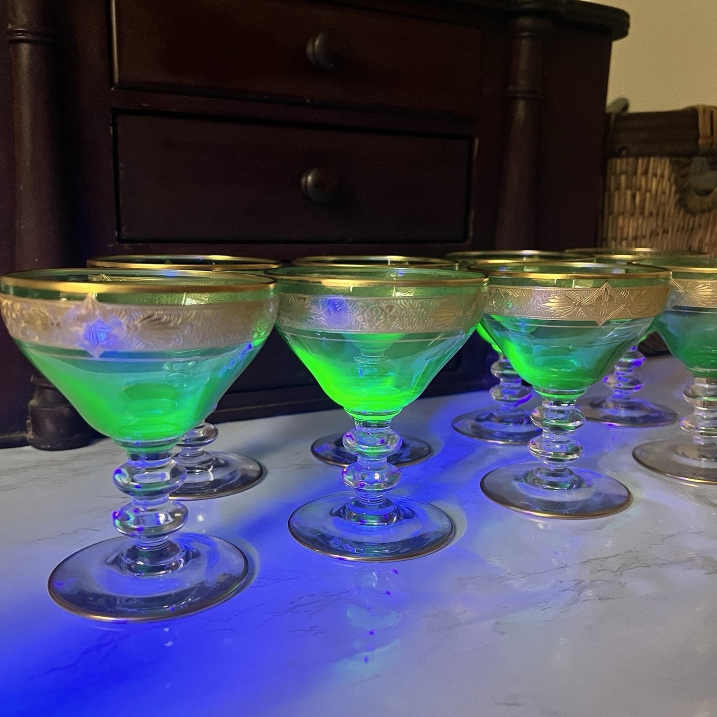 A set of clear stemmed cocktail glasses with gold rims, uv light shining on glasses, arranged on a white marble surface with blue lighting, with a dark wooden cabinet in the background.