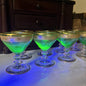 A set of clear stemmed cocktail glasses with gold rims, uv light shining on glasses, arranged on a white marble surface with blue lighting, with a dark wooden cabinet in the background.
