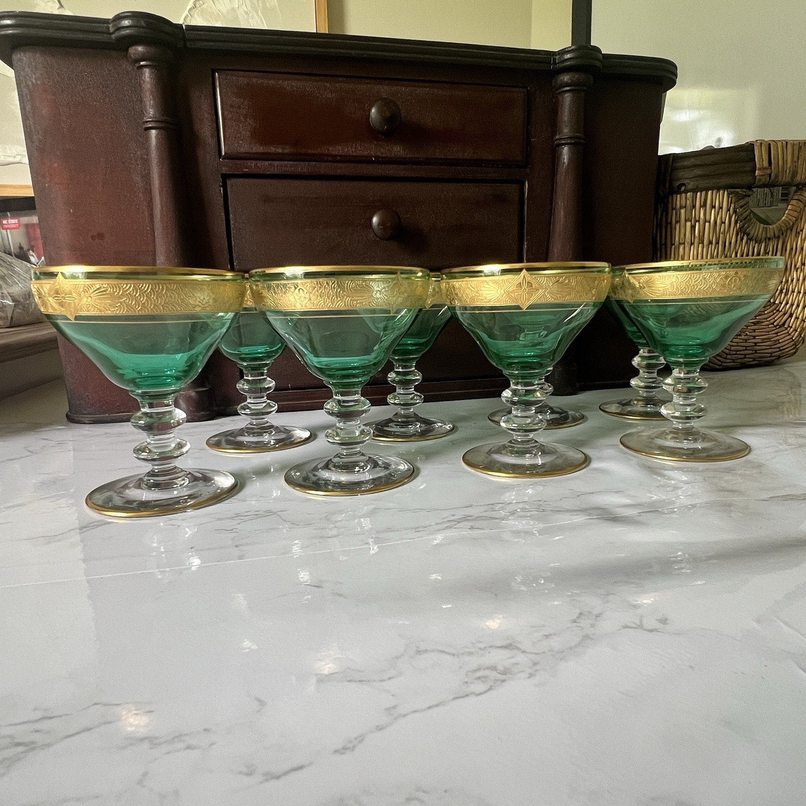 Six green coupe glasses with gold-trimmed rims arranged on a white marble surface in front of a dark wooden cabinet.