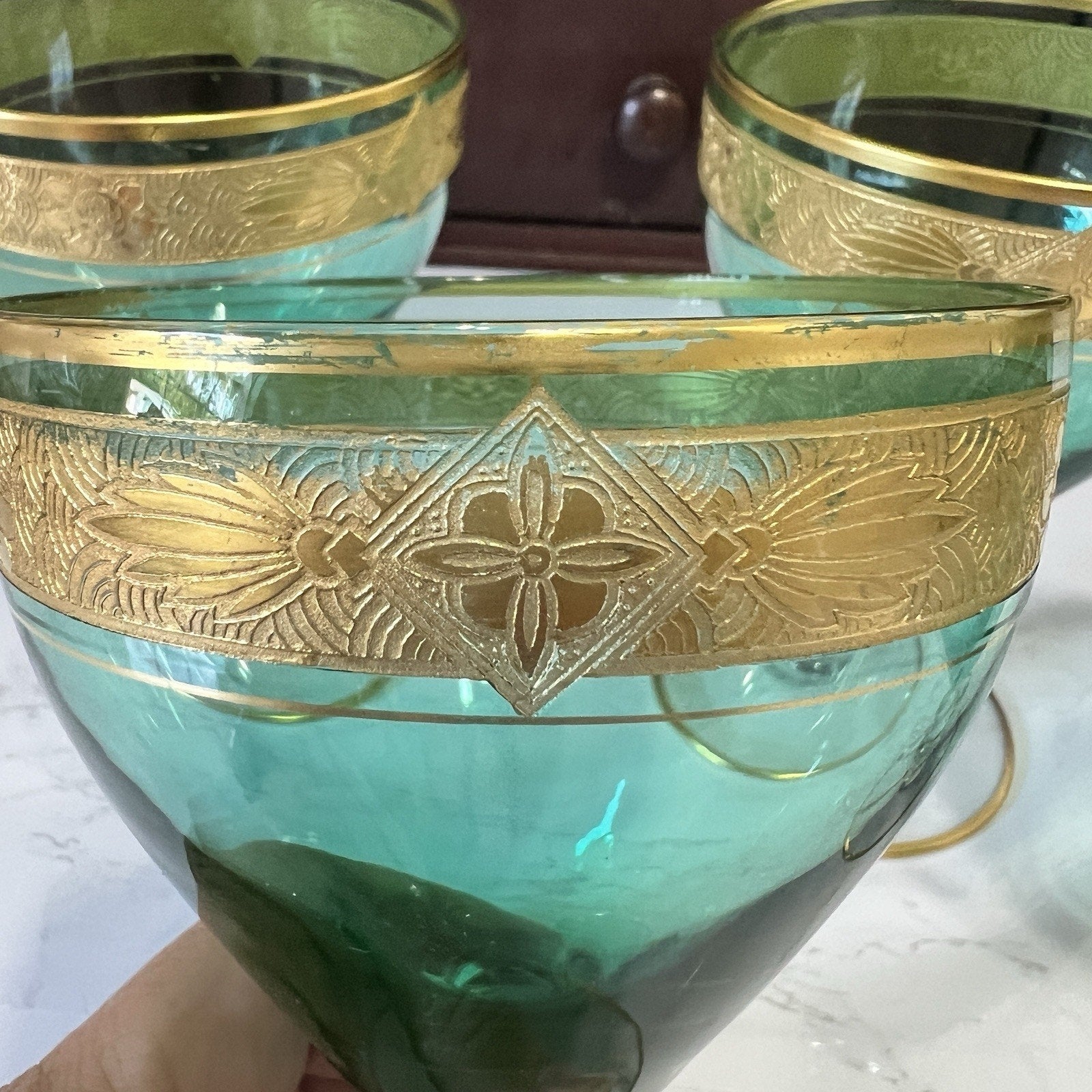 A close-up of a teal-green glass with an ornate gold band featuring a floral motif, held by a hand against a background of similar glassware.