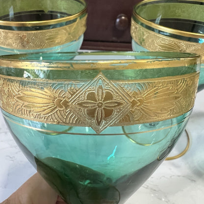 A close-up of a teal-green glass with an ornate gold band featuring a floral motif, held by a hand against a background of similar glassware.