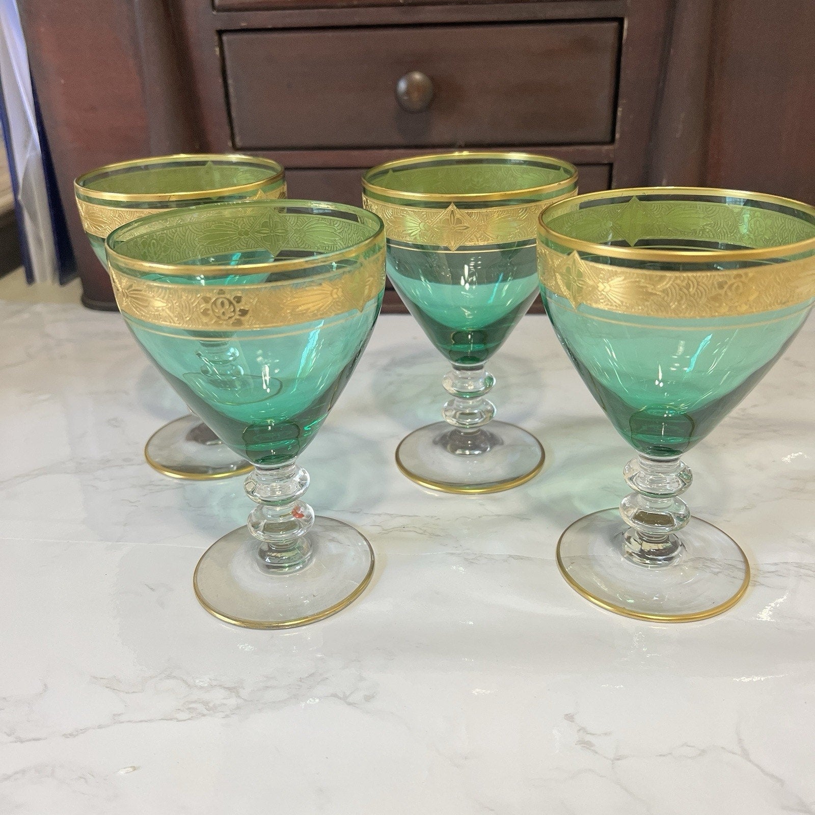 Four green and gold goblets arranged on a white marble surface with a dark wooden cabinet in the background.