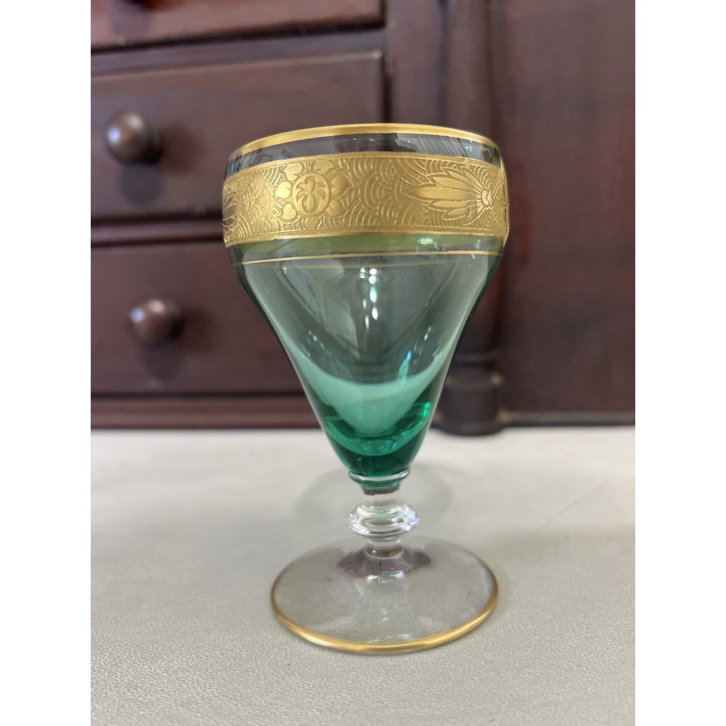 A vintage green glass goblet with ornate gold metal trim and a decorative floral band around the rim, set against a dark wooden cabinet background.