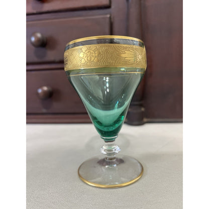 A vintage green glass goblet with ornate gold metal trim and a decorative floral band around the rim, set against a dark wooden cabinet background.