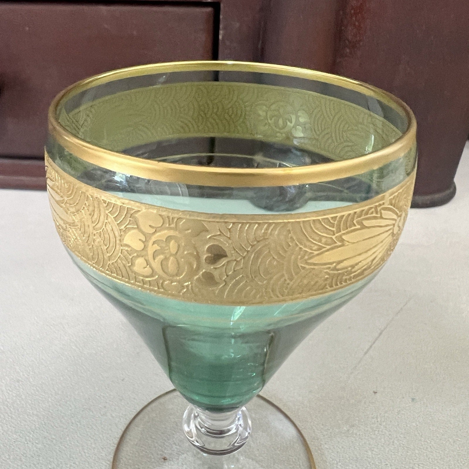 A green glass coupe-style cocktail glass featuring a wide gold band with embossed floral patterns and a gold rim, set against a neutral tabletop background.