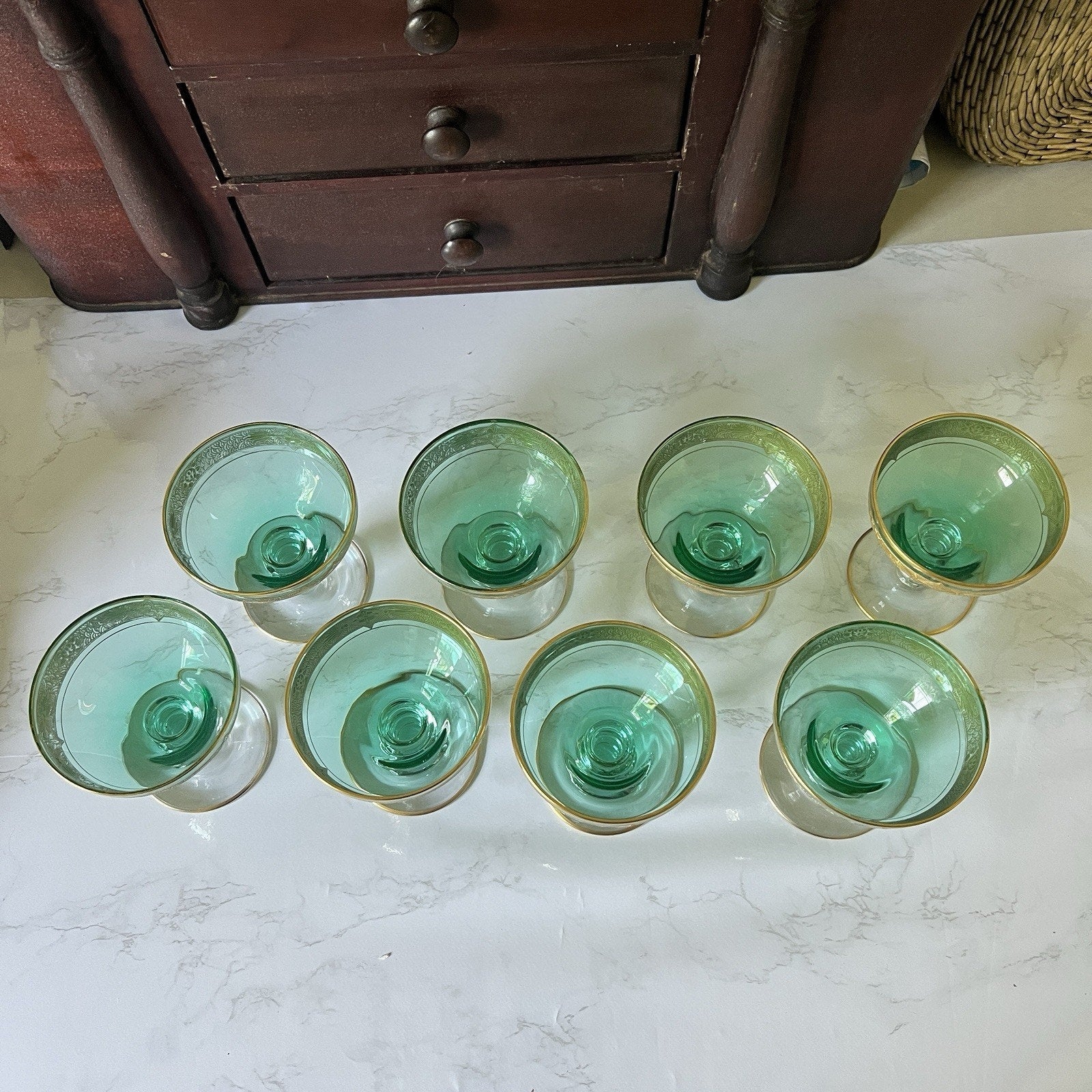 Eight small clear glasses with green interiors and gold rims, arranged on a white marble surface in front of a dark wooden drawer cabinet.