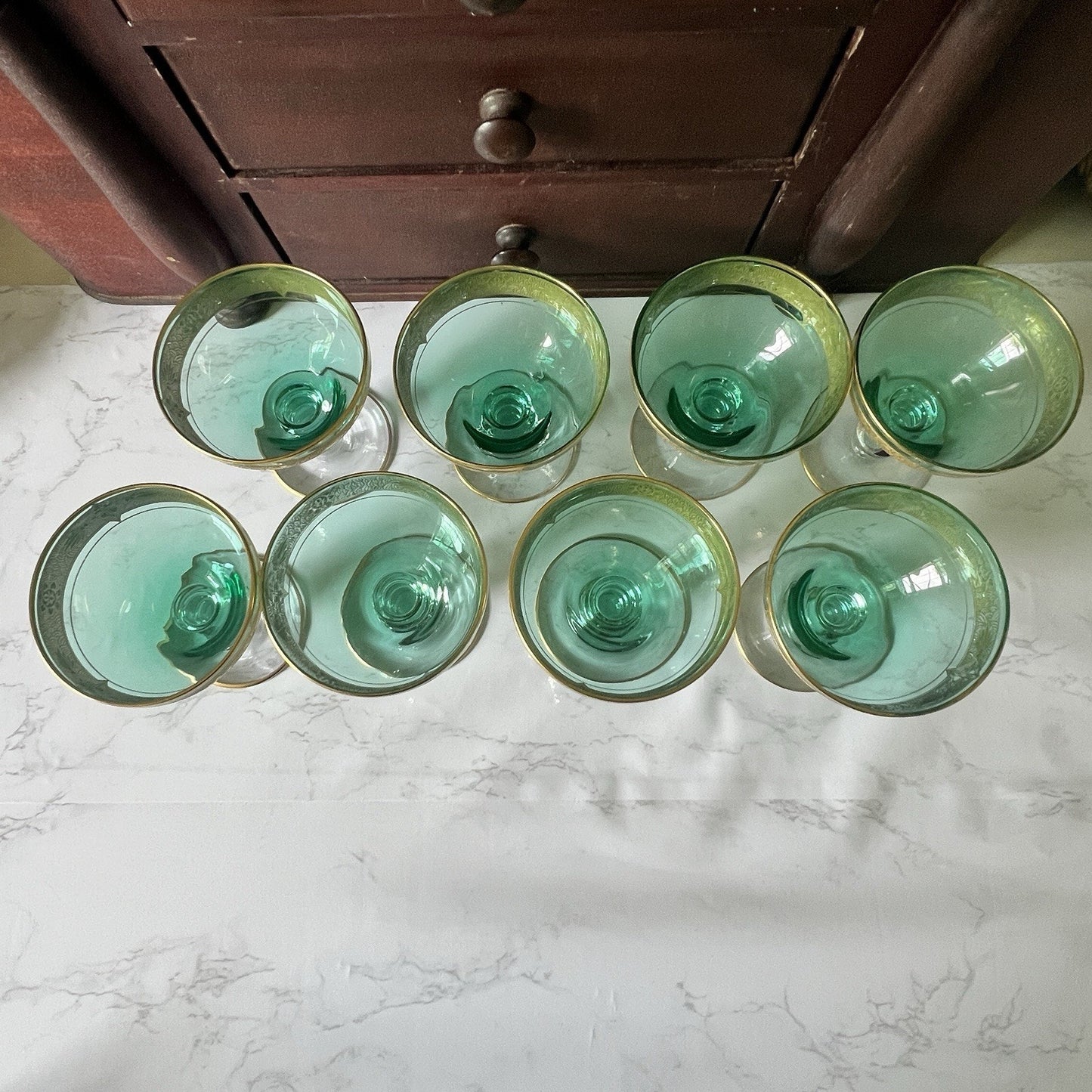 Six green-tinted glass tumblers arranged on a white marble-like surface with a dark wooden cabinet in the background.