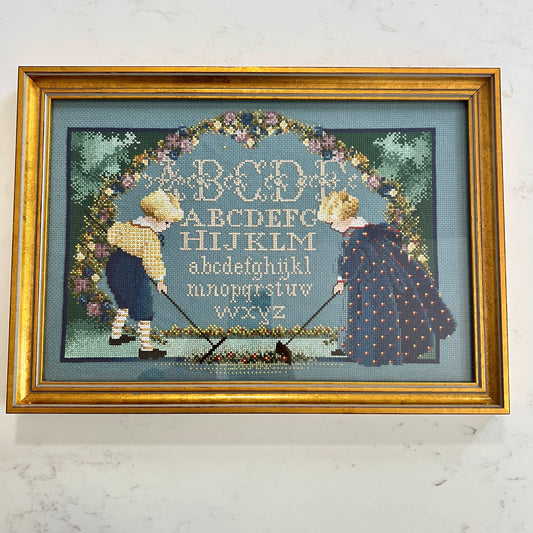 Framed cross-stitched picture of two children holding rakes with an alphabet design.