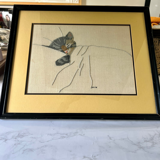 A framed art piece featuring a detailed embroidery of a sleeping cat under a blanket, set in a black frame with a wide beige mat, resting on a white marble surface.