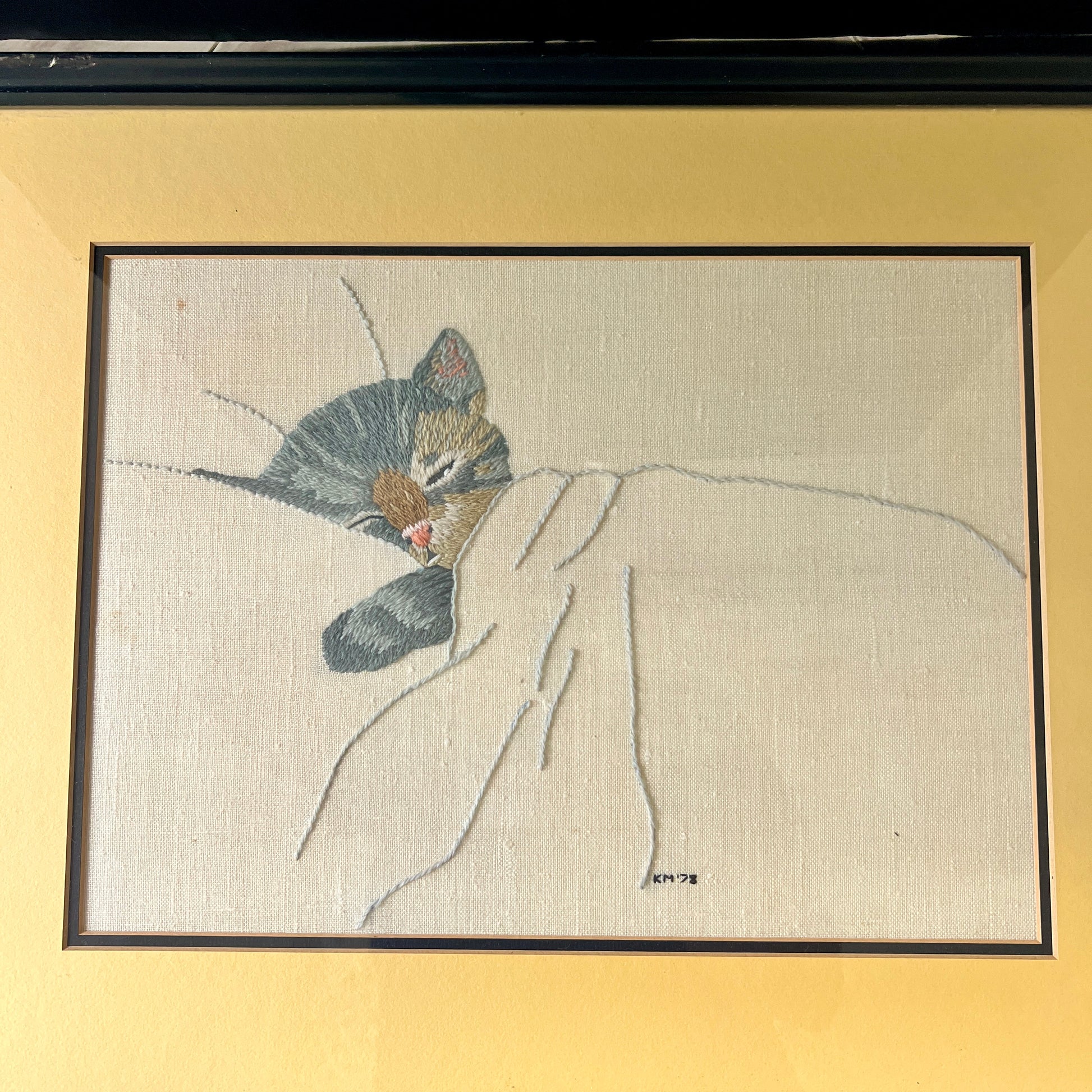 A framed textile artwork showing a sleeping cat nestled under a white blanket, mounted on a warm beige wall.