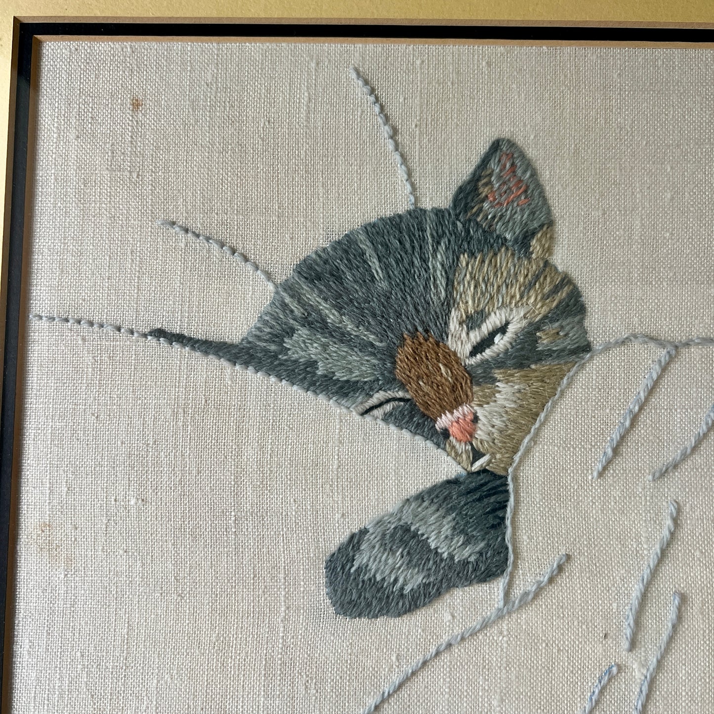 A close-up of framed needlepoint embroidery featuring a sleeping gray tabby kitten on a textured off-white canvas, with a dark brown wooden frame visible.