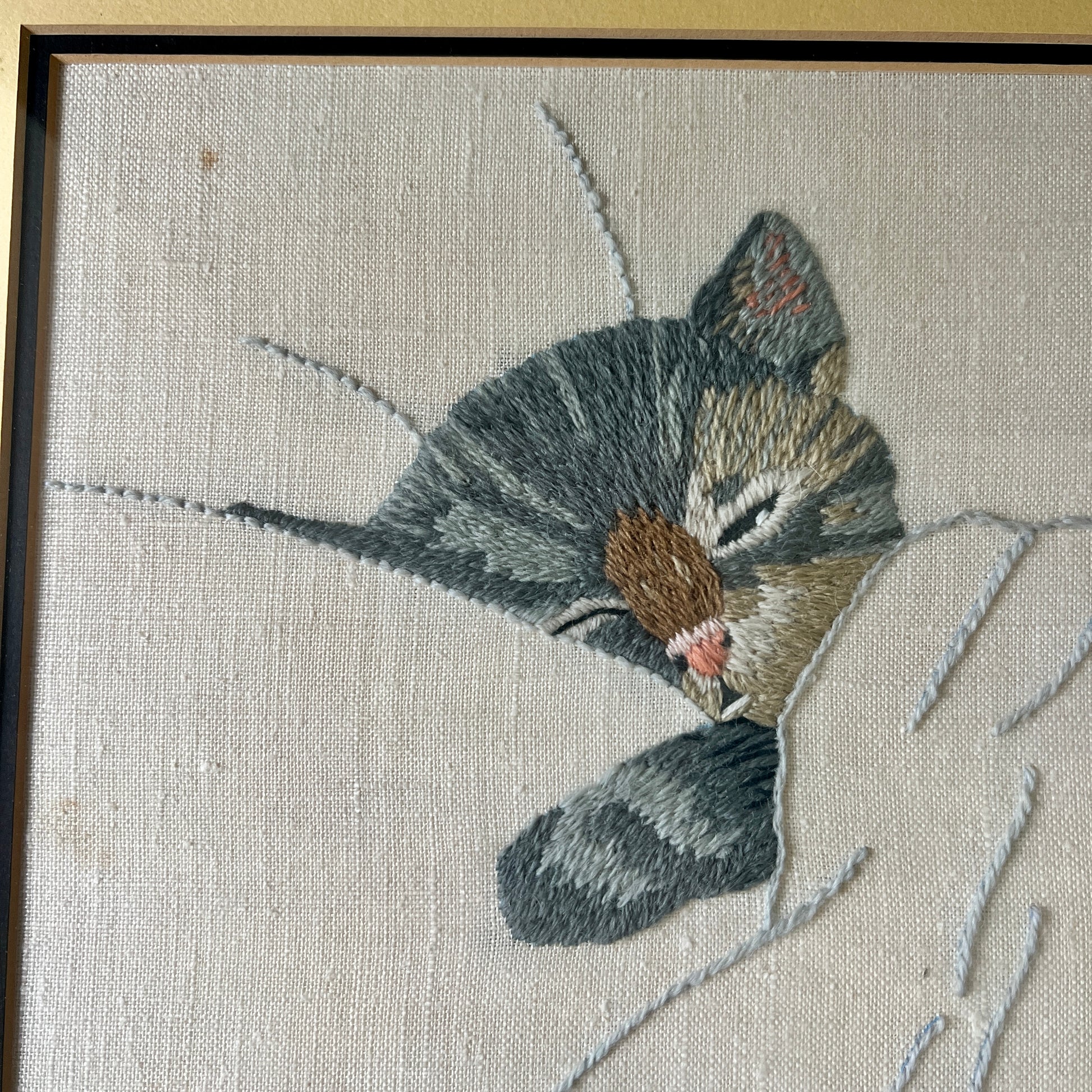 A close-up of framed needlepoint embroidery featuring a sleeping gray tabby kitten on a textured off-white canvas, with a dark brown wooden frame visible.