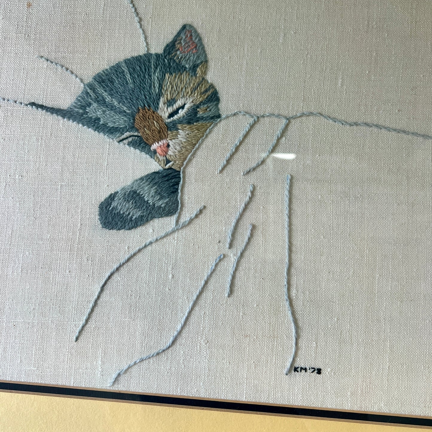A close-up of a framed textile artwork showing a sleeping gray tabby cat curled up under a white blanket, stitched on a light beige fabric background.