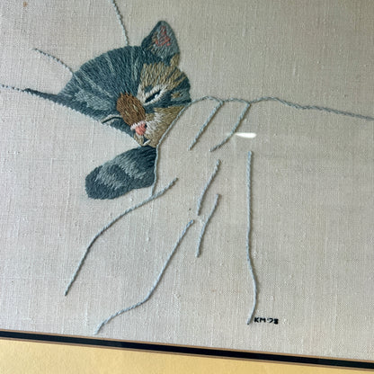 A close-up of a framed textile artwork showing a sleeping gray tabby cat curled up under a white blanket, stitched on a light beige fabric background.