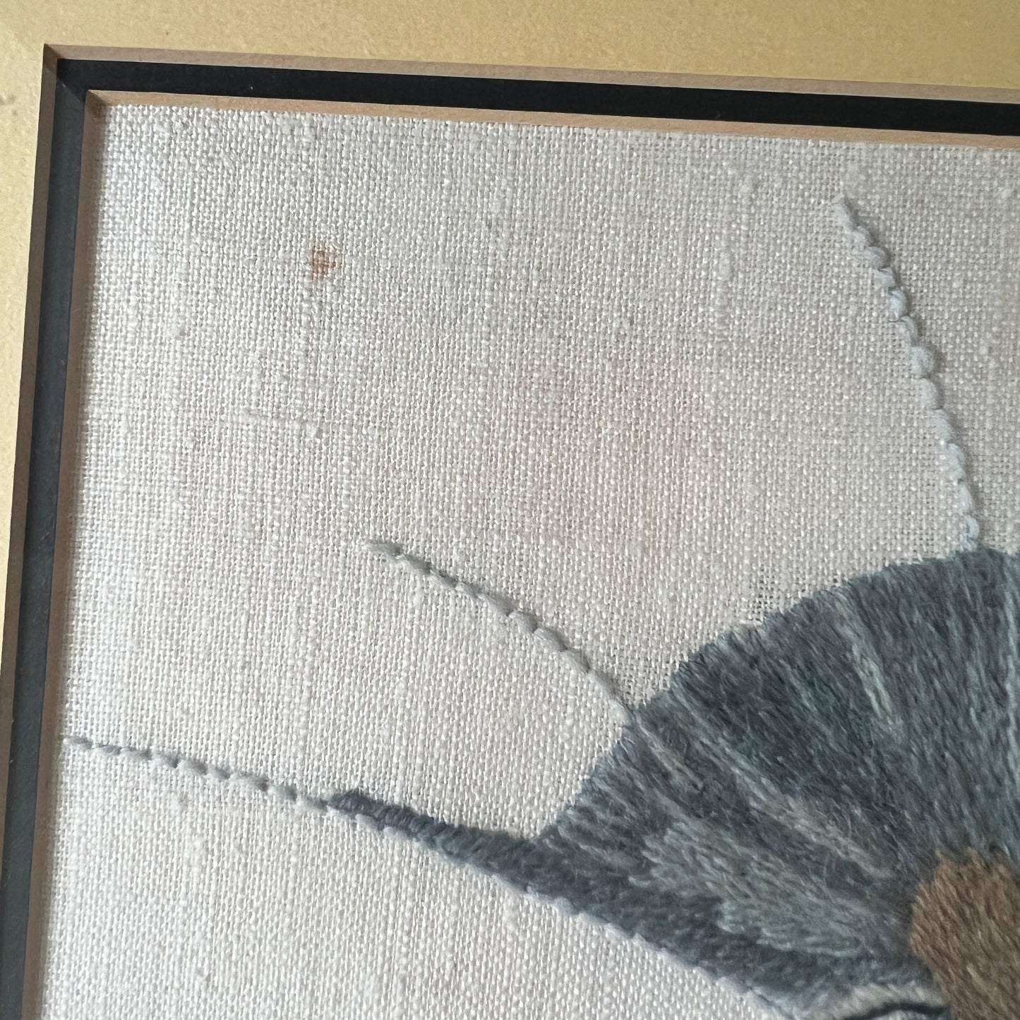Close-up of a framed textile artwork showing a light beige woven background, part of a gray embroidered shape, and a dark frame edge. showing a spot on canvas.