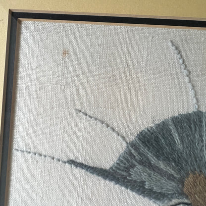 Close-up of a framed textile artwork showing a light beige woven background, part of a gray embroidered shape, and a dark frame edge. showing a spot on canvas.