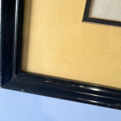 Close-up of a framed piece of wall art showing a glossy black frame, scuff marks on frame, textured beige mat, and a portion of a white woven fabric insert.