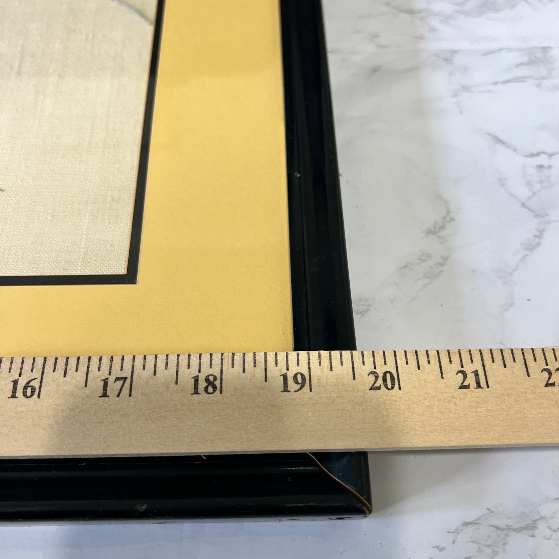 Close-up of a black picture frame with a wide gold mat, shown on a white marble surface with a metal ruler across the bottom to indicate size.