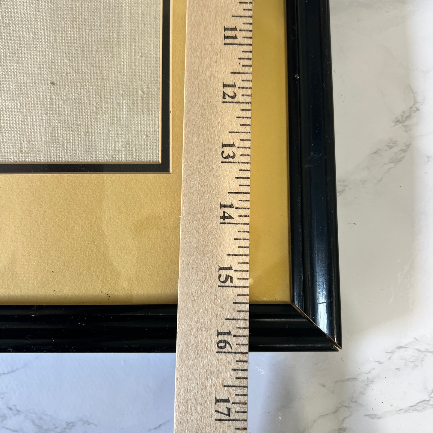 Close-up of a black picture frame with a gold mat and a wooden ruler showing the dimensions, set against a white marble surface.