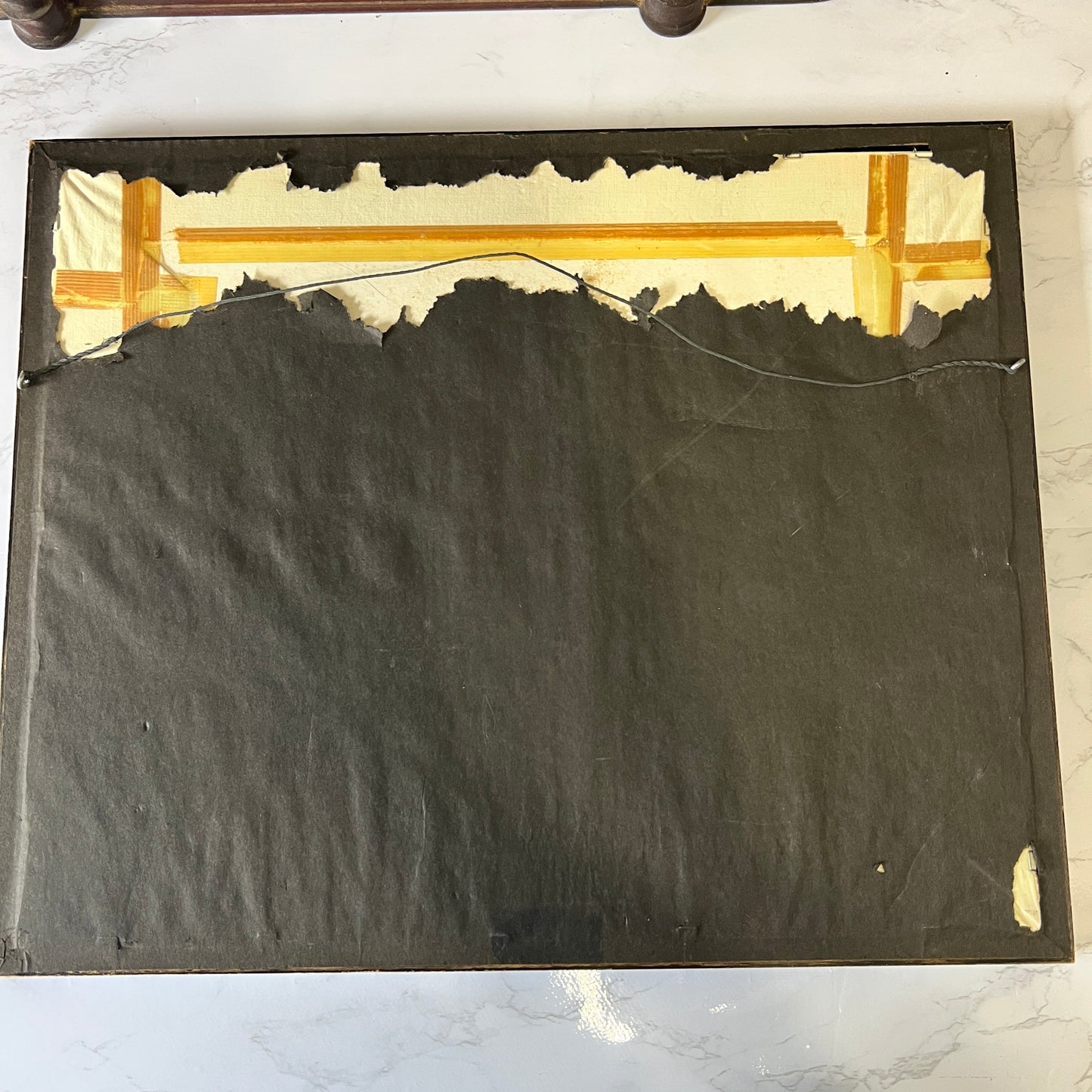 The back side of a framed artwork or panel, showing a dark charcoal fabric backing with torn off cream paper along the top edge and a piece of tape near the upper right corner, placed on a light marble surface.