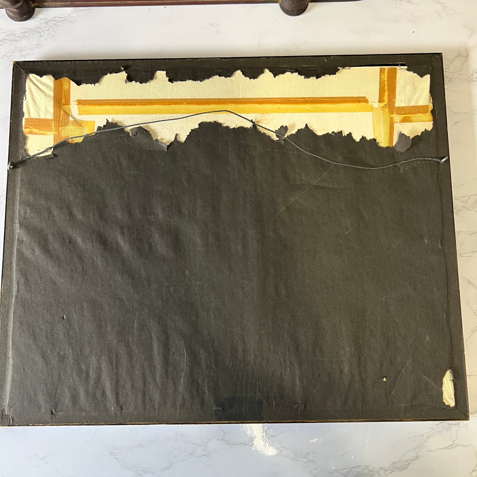 The back side of a framed artwork or panel, showing a dark charcoal fabric backing with torn off cream paper along the top edge and a piece of tape near the upper right corner, placed on a light marble surface.
