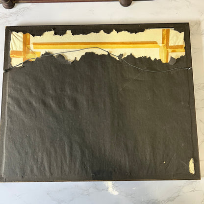 The back side of a framed artwork or panel, showing a dark charcoal fabric backing with torn off cream paper along the top edge and a piece of tape near the upper right corner, placed on a light marble surface.