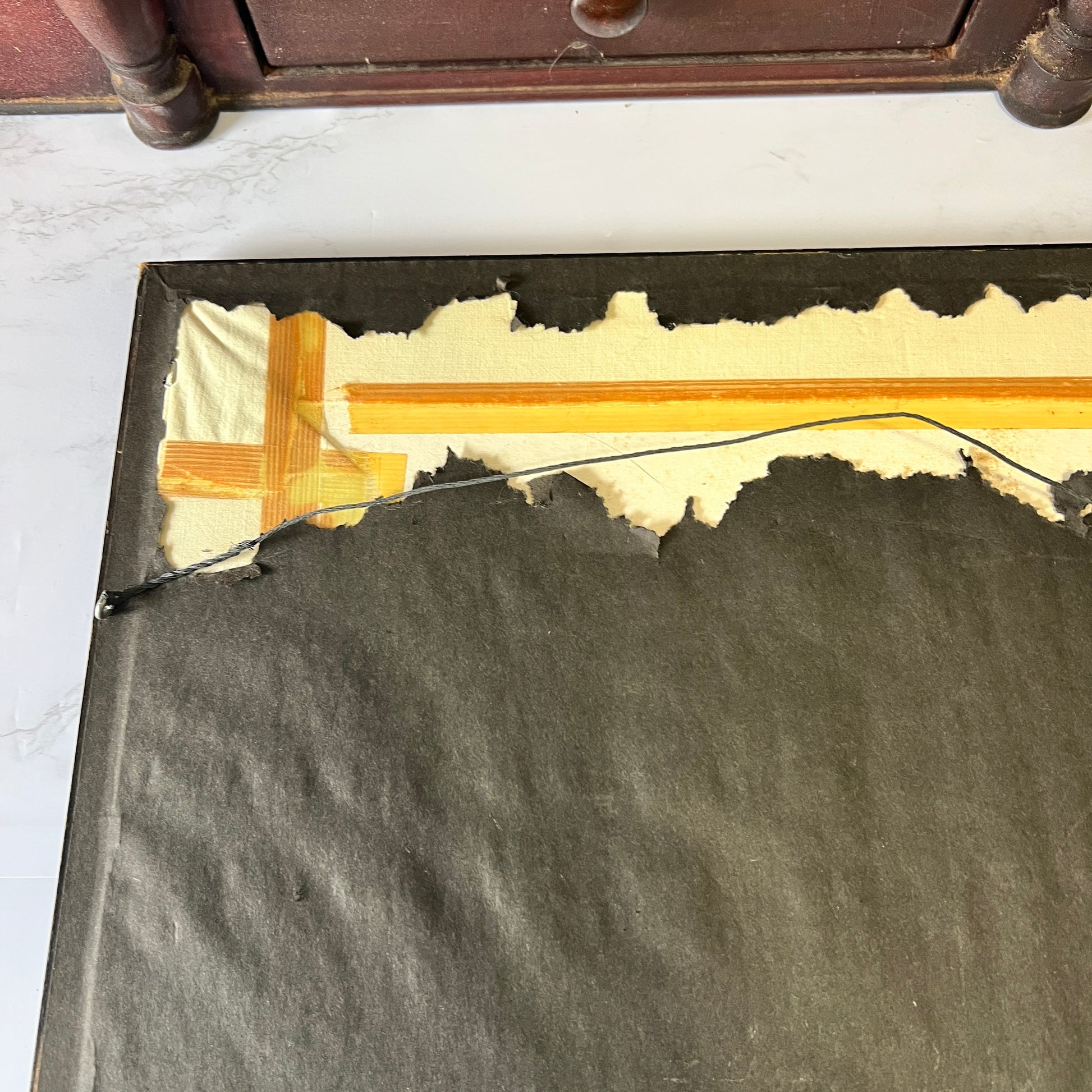 A close-up of the back of a dark gray or black rectangular mat, showing a torn edge with beige backing material and a yellow-orange adhesive strip applied to a white marble surface.
