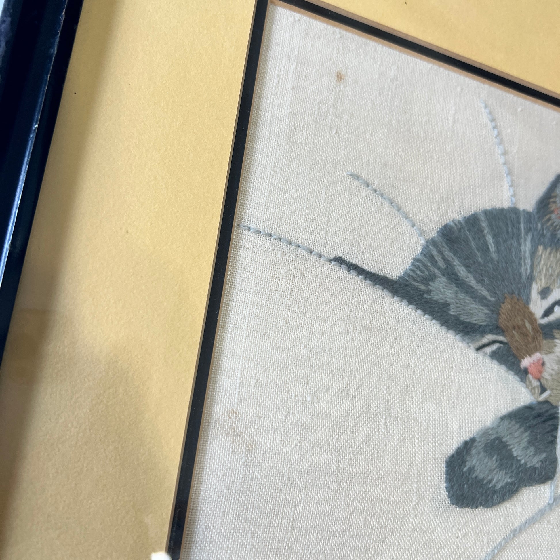 Close-up of a framed needlepoint canvas featuring a gray tabby cat, with a beige mat and a black frame visible.