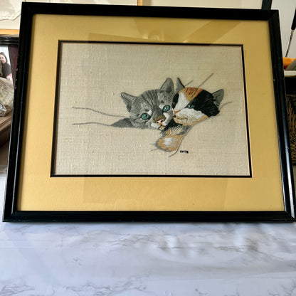 A framed needlepoint canvas featuring a gray tabby cat and a calico cat, presented in a black frame with a wide beige mat on a marble surface.