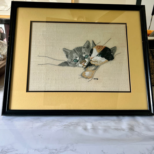 A framed needlepoint canvas featuring a gray tabby cat and a calico cat, presented in a black frame with a wide beige mat on a marble surface.
