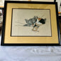 A framed needlepoint canvas featuring a gray tabby cat and a calico cat, presented in a black frame with a wide beige mat on a marble surface.