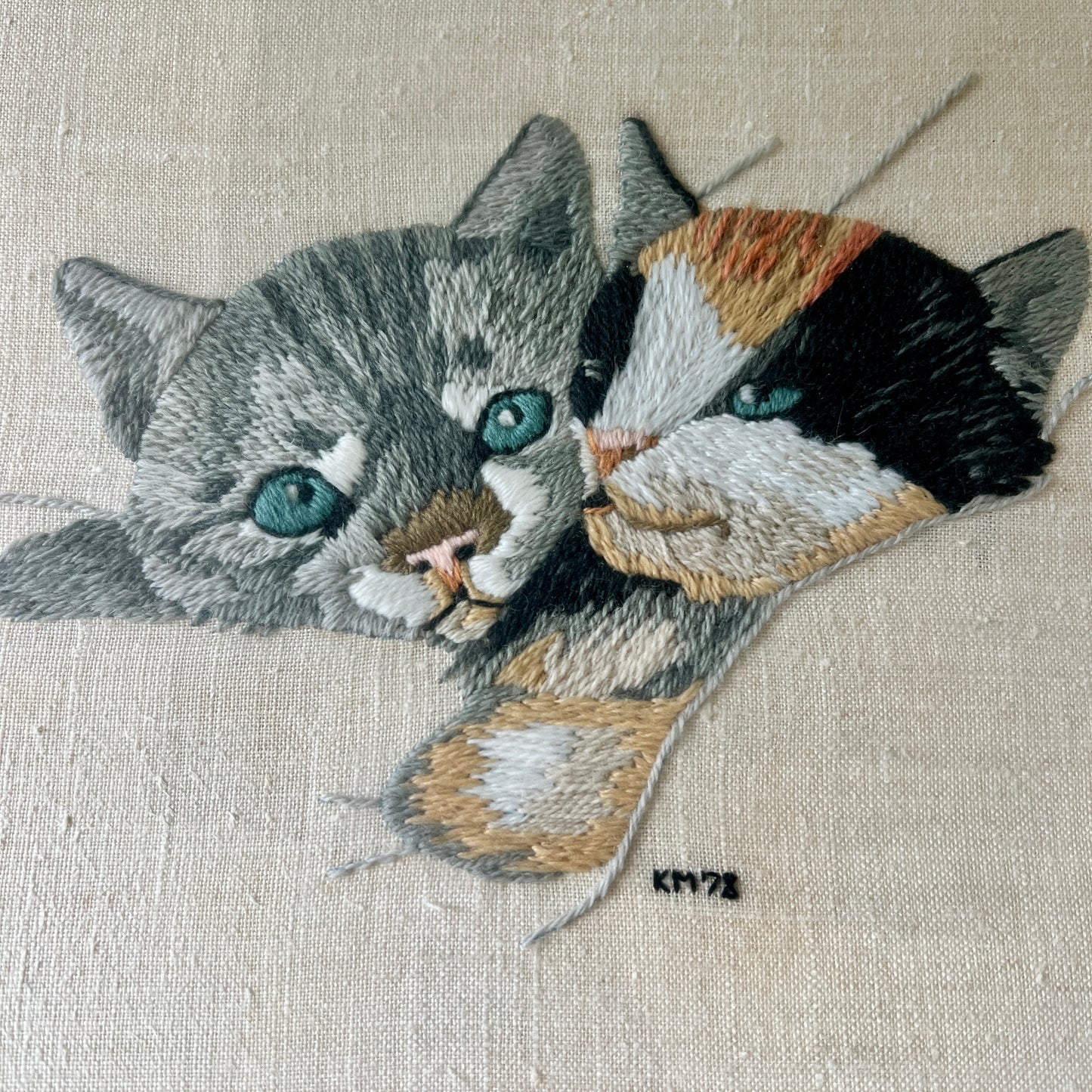 A close-up of a hand-embroidered textile featuring two cats, one gray tabby and one black, white, and orange calico, with blue-green eyes, on a beige linen background.