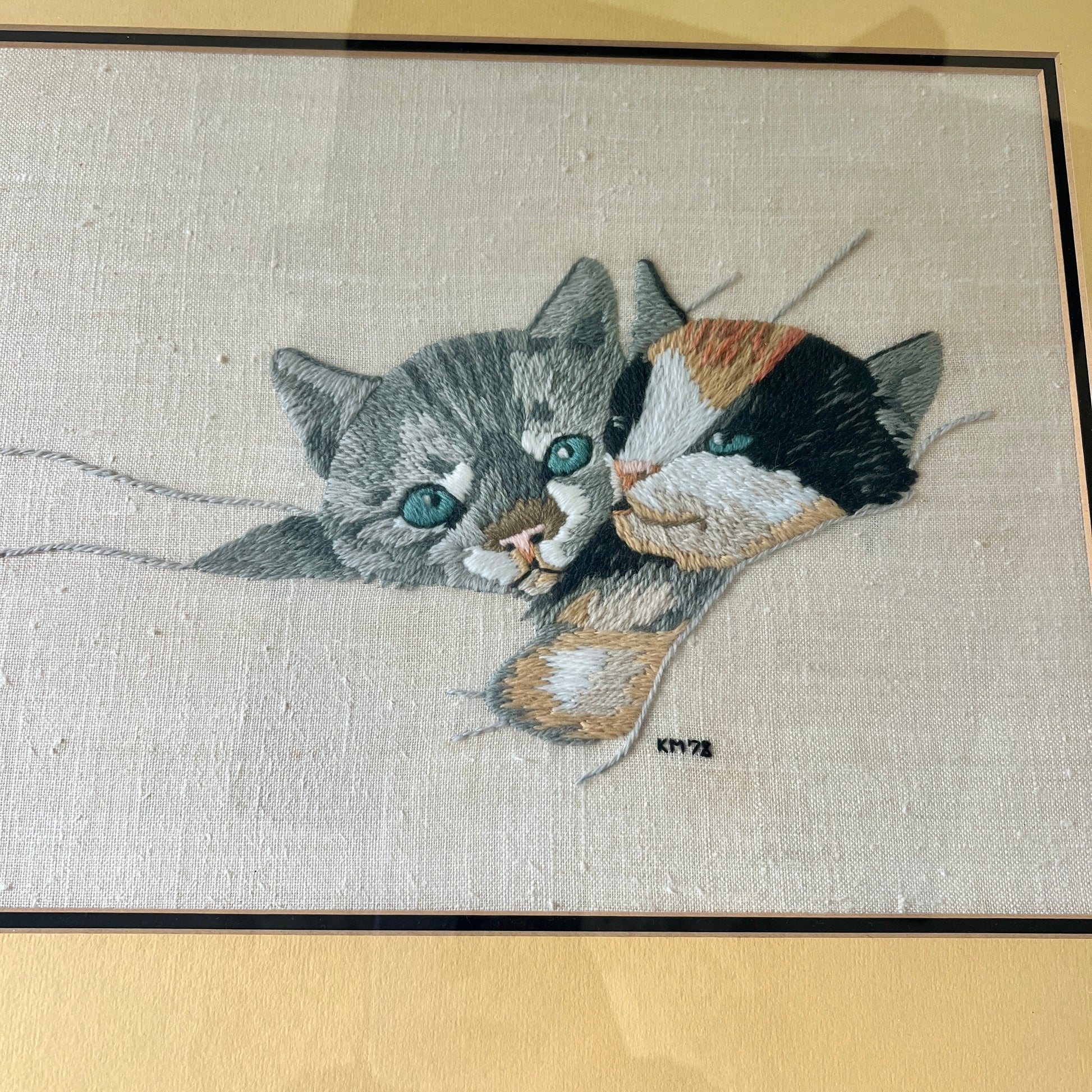A close-up of a hand-embroidered textile featuring two cats, one gray tabby and one black, white, and orange calico, with blue-green eyes, on a beige linen background with yellow mat.