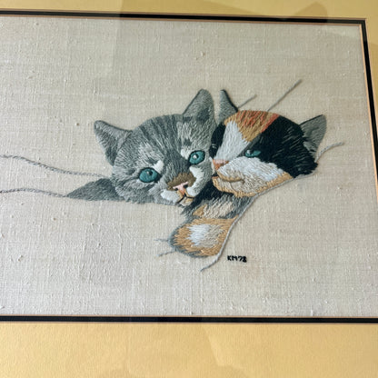 A close-up of a hand-embroidered textile featuring two cats, one gray tabby and one black, white, and orange calico, with blue-green eyes, on a beige linen background with yellow mat.
