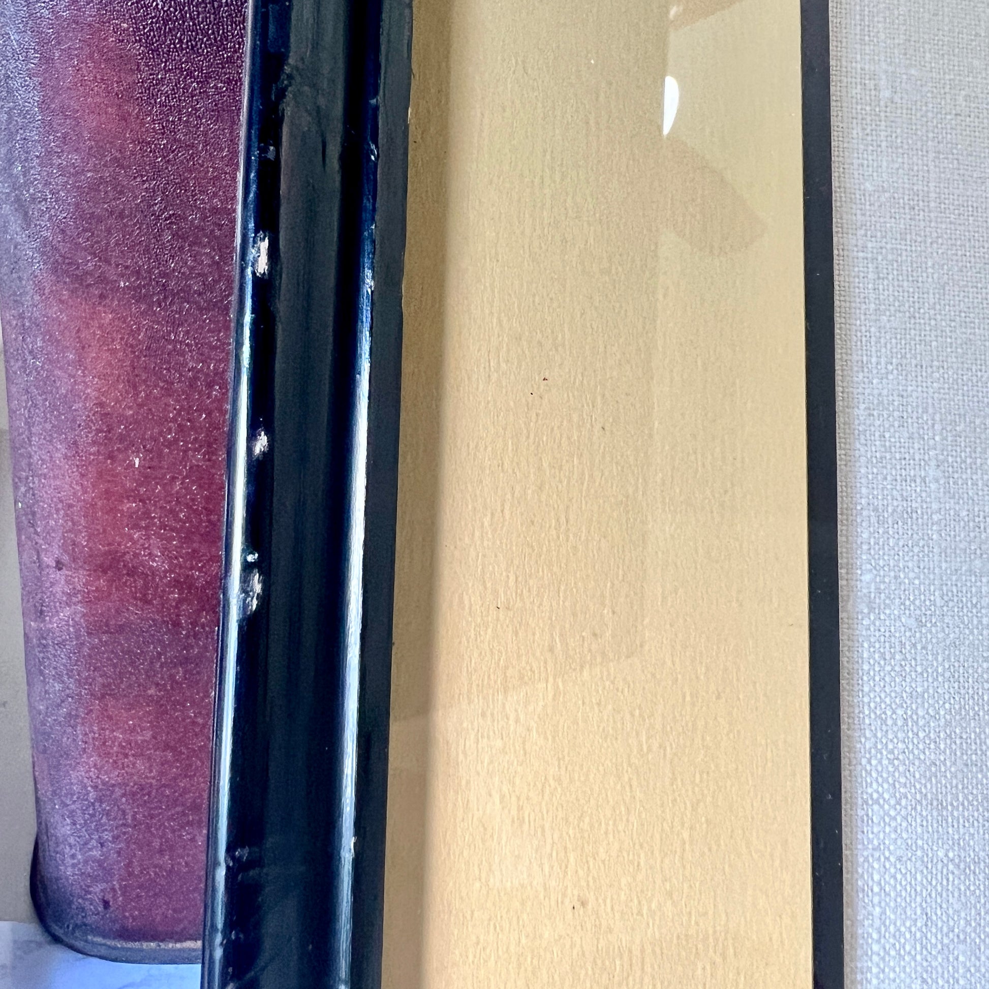 Close-up of a framed art print or panel with a black frame, a beige/tan textured center, and a reddish-purple textured vertical element on the left.