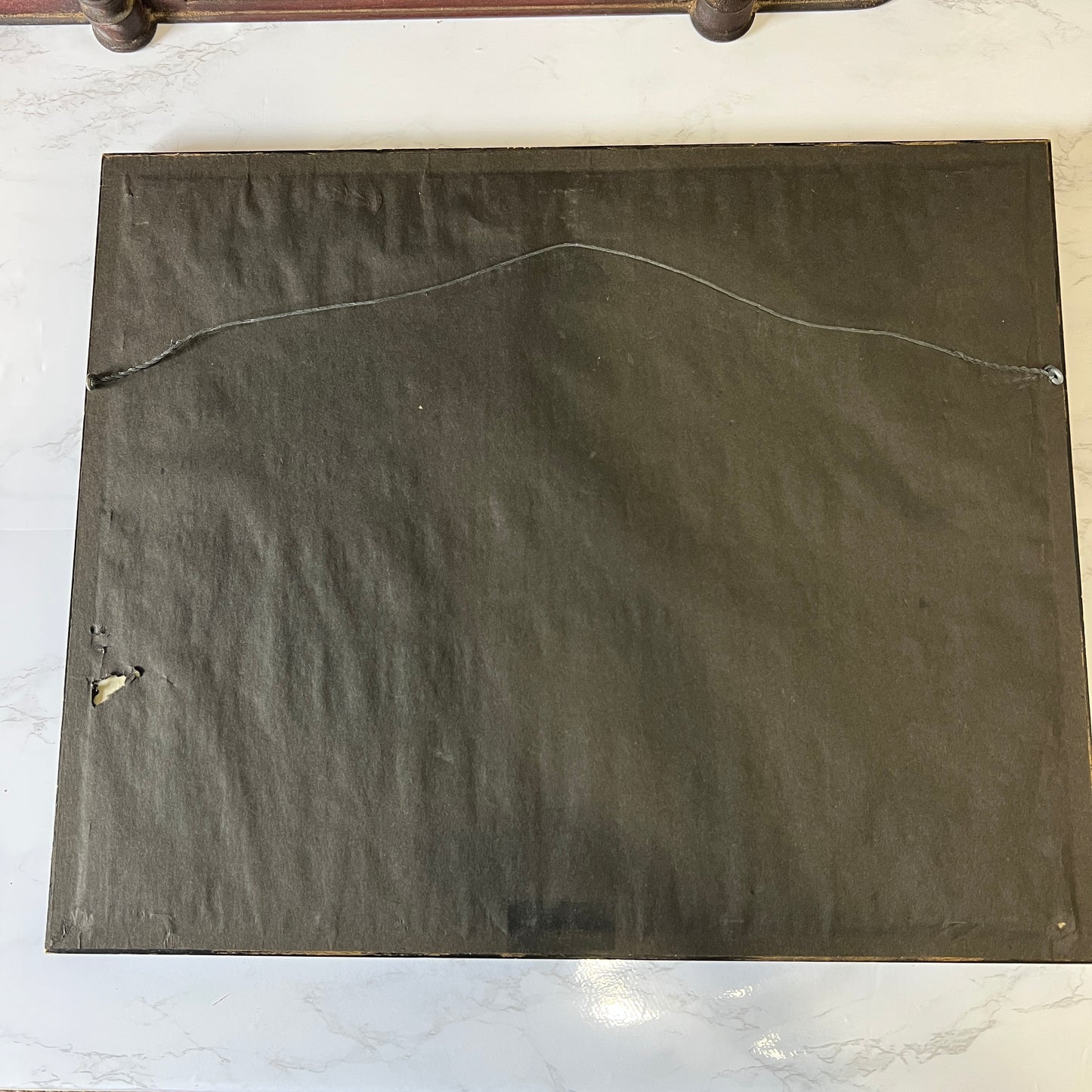 The back of a square dark gray or charcoal-colored framed wall art piece, showing the mounting canvas texture and a metal hanging wire, photographed on a white marble surface.