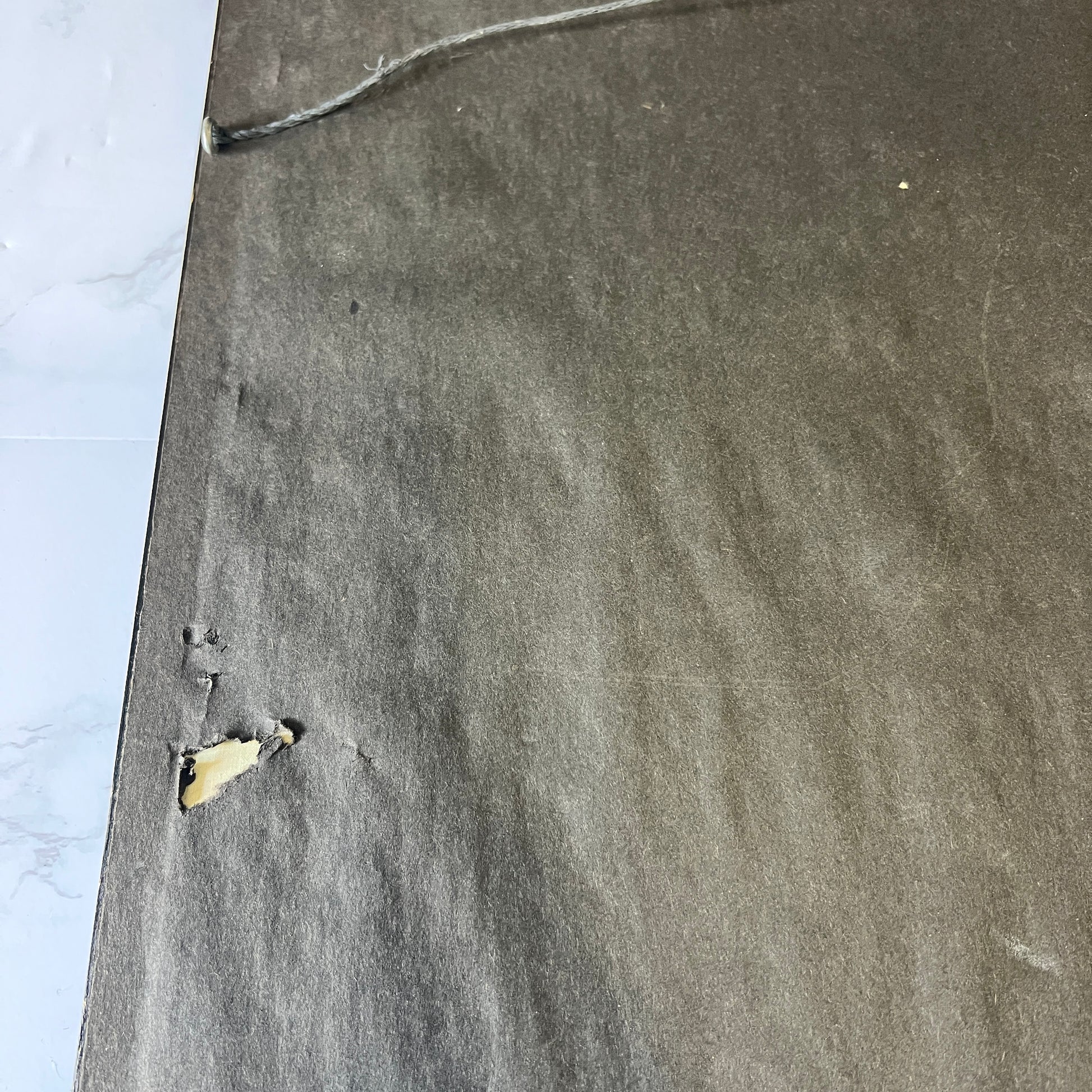 A close-up of a dark gray textured carpet or rug with a small torn section showing a beige backing, placed next to a white marble-patterned surface.