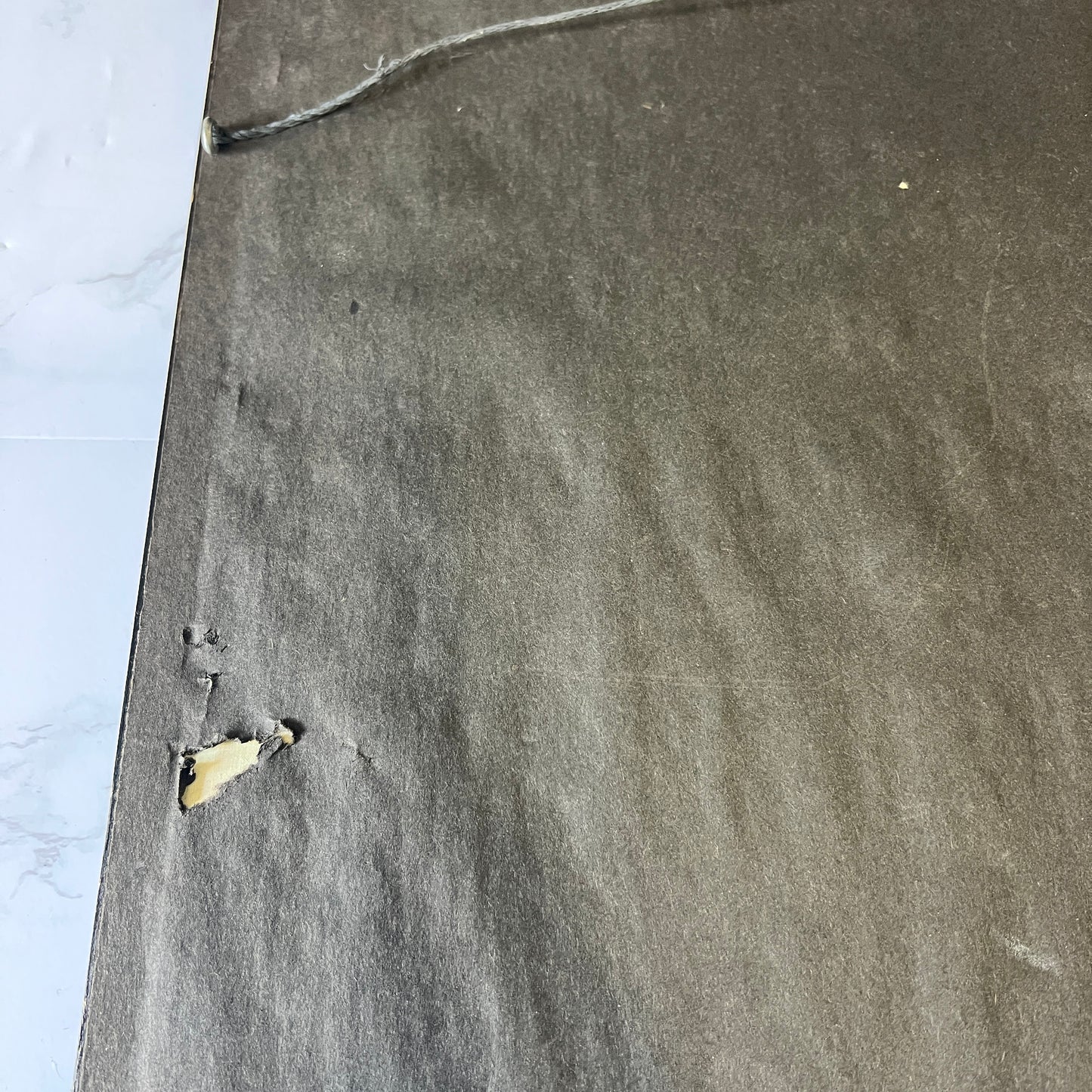 A close-up of a dark gray textured carpet or rug with a small torn section showing a beige backing, placed next to a white marble-patterned surface.