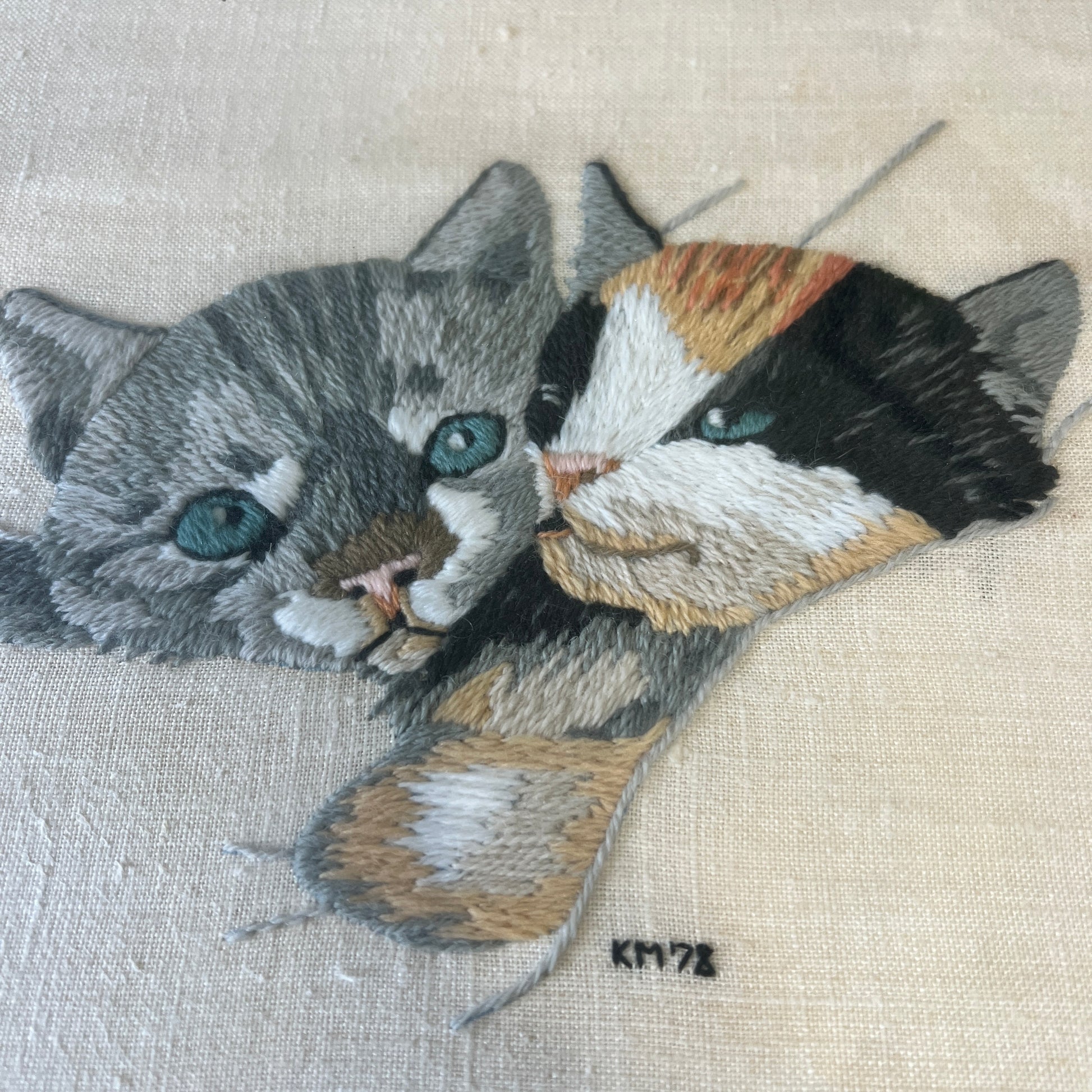 Close-up of a detailed embroidered cat portrait on light beige fabric, showing two cats with blue-green eyes, one gray and one black/white/tan, with a small artist signature 'KM78' below.