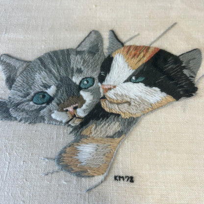 Close-up of a detailed embroidered cat portrait on light beige fabric, showing two cats with blue-green eyes, one gray and one black/white/tan, with a small artist signature 'KM78' below.